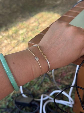 Sunlit outdoor wrist wearing a green jade bangle and dainty gold and silver bracelet stack with a small letter charm, resting on a wooden patio table with blurred grass and cords in the background.