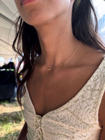 Close-up of a woman's neck and collarbone wearing a delicate gold cross necklace paired with a white lace dress in a sunlit outdoor setting