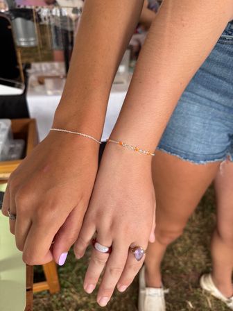 Two wrists side-by-side wearing delicate silver chain bracelets—one with tiny orange beads—paired with rings and lavender nail polish, set against denim shorts and a grassy outdoor craft market backdrop.