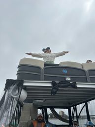 Person standing on the upper deck of a pontoon boat with arms outstretched against an overcast sky, other passengers seated below and water and distant shoreline buildings visible.