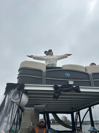 Person standing on the upper deck of a pontoon boat with arms outstretched against an overcast sky, other passengers seated below and water and distant shoreline buildings visible.