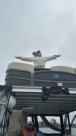 Person standing on the upper deck of a pontoon boat with arms outstretched against an overcast sky, other passengers seated below and water and distant shoreline buildings visible.