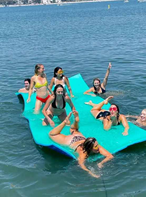 Group of friends lounging and playing on a large turquoise floating mat in calm bay waters near a shoreline and boats on a sunny summer day.