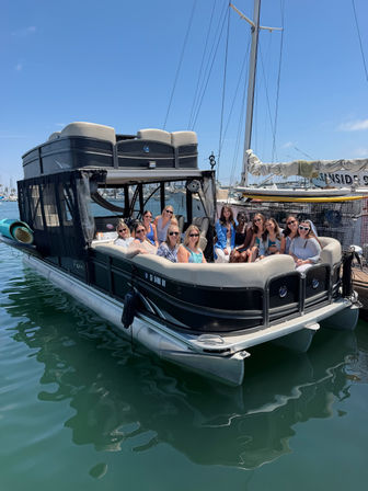 Group of friends smiling on a black pontoon boat docked at a sunny marina with sailboats and tall masts in the background and calm green harbor water reflecting the hull.