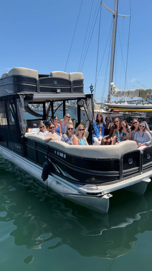 Group of friends smiling on a black pontoon boat docked at a sunny marina with sailboats and tall masts in the background and calm green harbor water reflecting the hull.