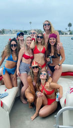 Group of eight women in colorful bikinis smiling and posing on a pontoon boat, some holding canned drinks, with palm trees and shoreline houses in the background under a cloudy sky — beach boat party vibe.
