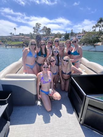 Smiling group of young women in colorful bikinis posing on a pontoon boat in a sunny coastal bay, with palm-lined waterfront homes and blue skies in the background.