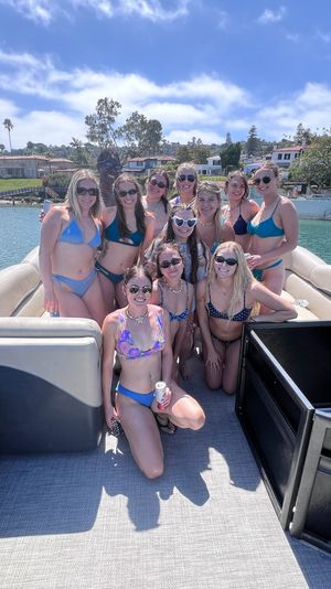 Smiling group of young women in colorful bikinis posing on a pontoon boat in a sunny coastal bay, with palm-lined waterfront homes and blue skies in the background.