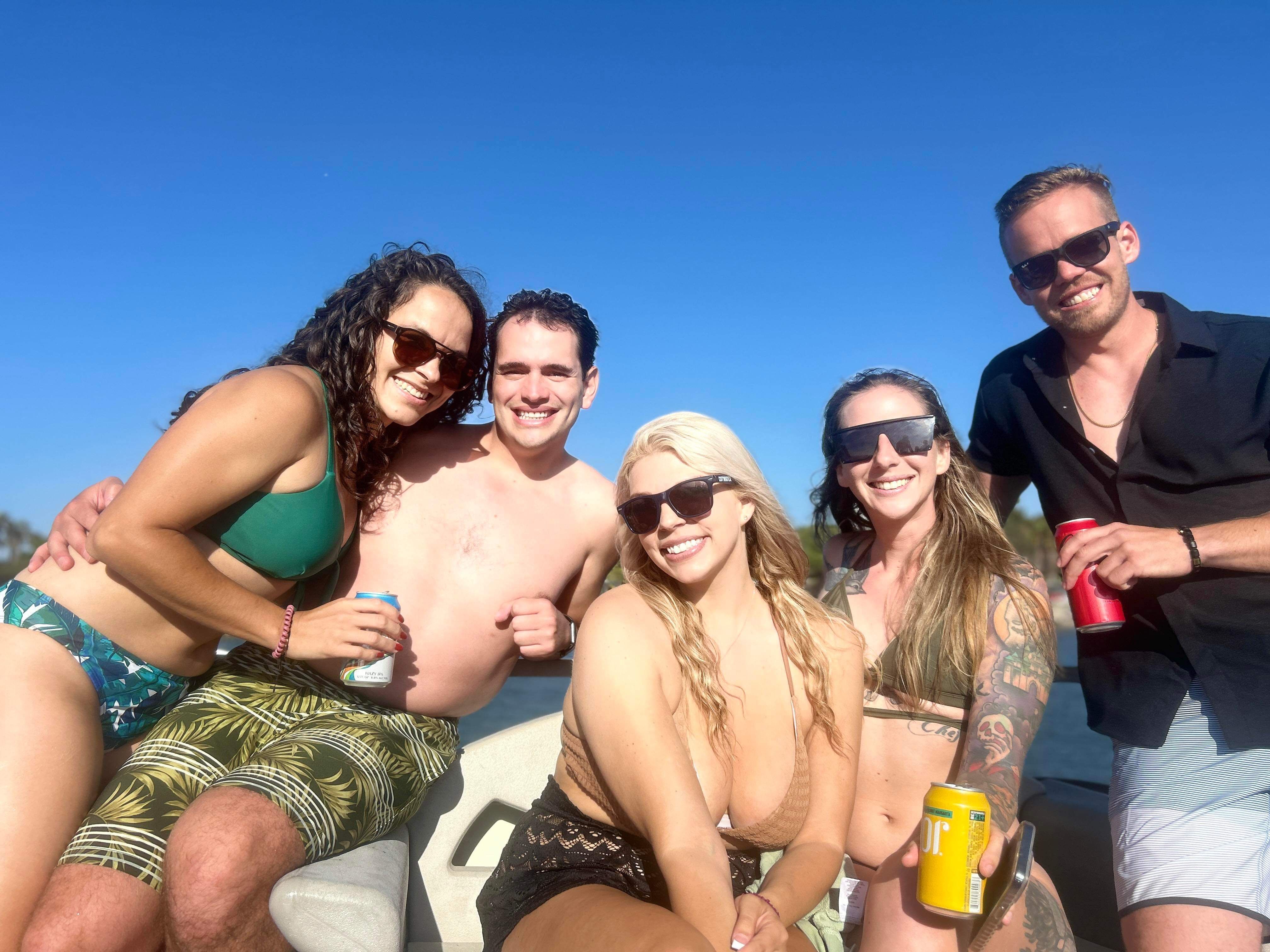 Five friends in swimsuits and sunglasses on a boat, holding canned drinks and smiling under a clear blue sky on a sunny lake day.