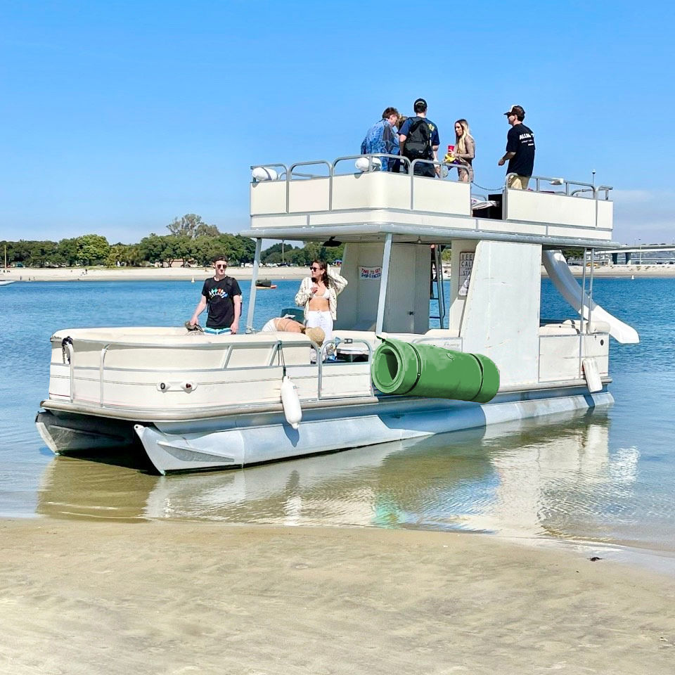 Group of people on a two-level pontoon boat beached in shallow coastal water on a sunny day, with a slide and green float attached and a sandy shoreline with trees in the background.