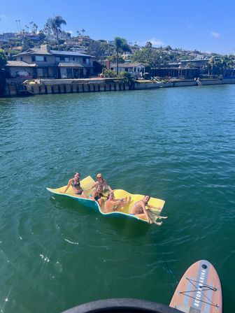 Group of friends lounging on a yellow floating mat in blue-green bay water with hillside waterfront homes and a paddleboard visible in the foreground on a sunny day