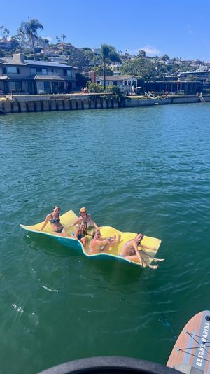 Group of friends lounging on a yellow floating mat in blue-green bay water with hillside waterfront homes and a paddleboard visible in the foreground on a sunny day