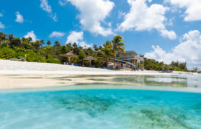 Sun-drenched tropical white-sand beach with crystal turquoise water, palm trees, thatched umbrellas and a raised beachfront house under a bright blue sky.