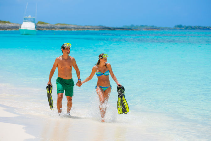 Couple holding hands walking from turquoise water onto a white-sand tropical beach, carrying yellow-green snorkel masks and fins with a boat anchored in the distance.