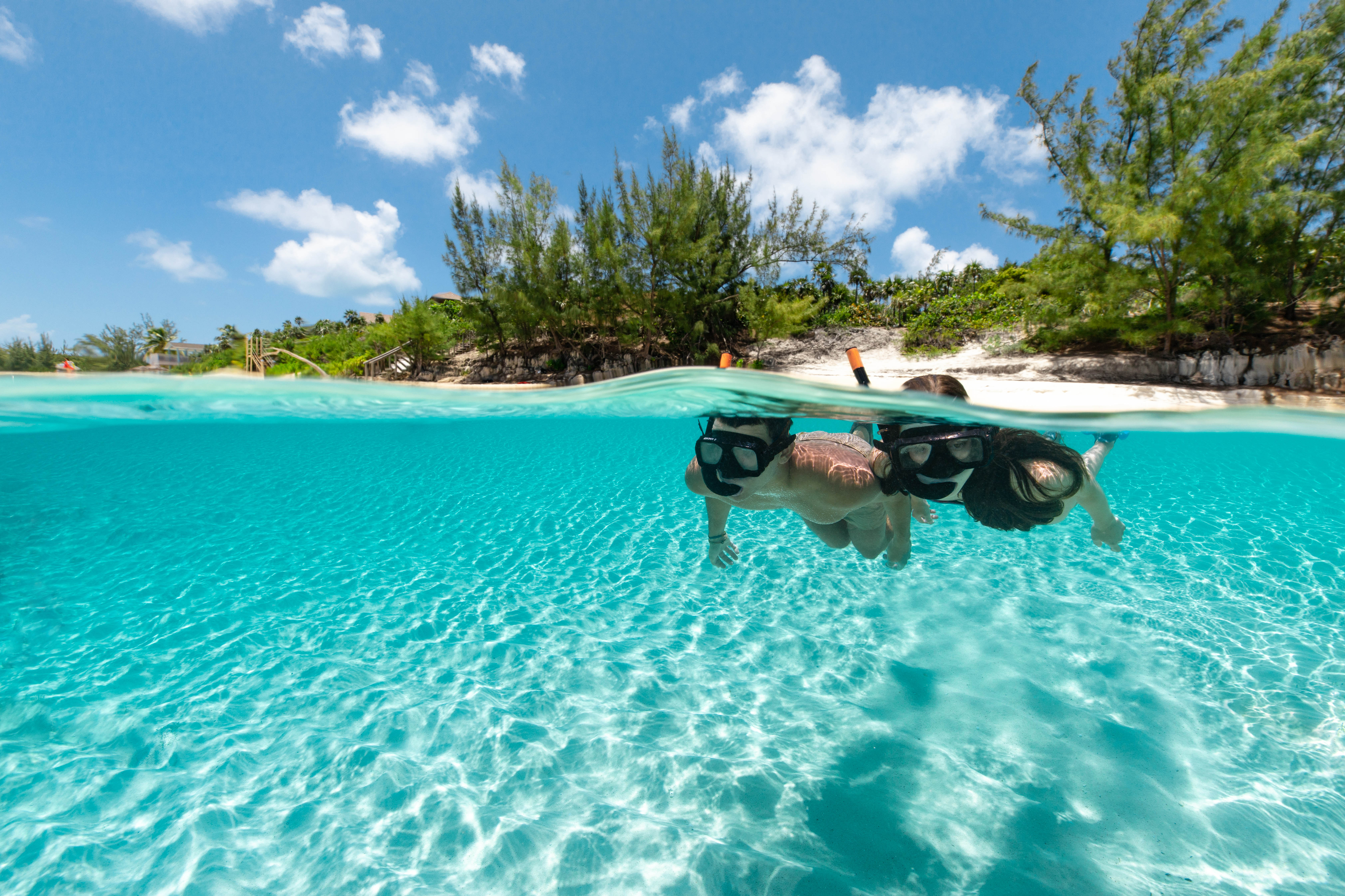 Over-under shot of two snorkelers in crystal-clear turquoise water above a white-sand tropical beach with green coastal trees and a bright blue sky.