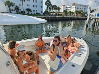 Smiling friends in colorful swimsuits lounging on a white motorboat at a sunny marina, palm trees and waterfront condos reflecting in calm water.