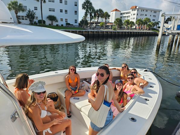 Smiling friends in colorful swimsuits lounging on a white motorboat at a sunny marina, palm trees and waterfront condos reflecting in calm water.