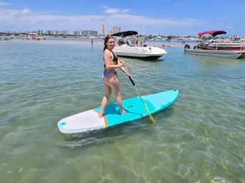 Person paddleboarding on a turquoise stand-up paddleboard in clear shallow coastal water near a sandy sandbar with anchored boats and a beachfront high-rise skyline on a sunny day.