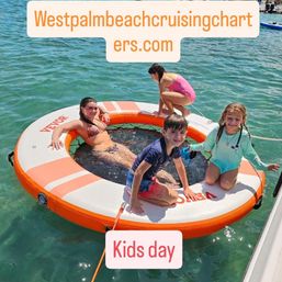 Smiling kids playing on an orange circular inflatable swim platform in clear turquoise West Palm Beach waters beside a boat for a sunny kids day outing.