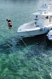 Two girls mid-air jumping off a white center-console boat into clear turquoise water beside a moored vessel with people on deck and a striped towel