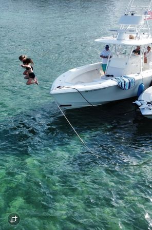 Two girls mid-air jumping off a white center-console boat into clear turquoise water beside a moored vessel with people on deck and a striped towel