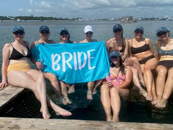 Bachelorette party at a coastal marina: eight women in swimsuits and matching caps sit on a wooden dock with their feet in the water, holding a bright turquoise banner reading BRIDE under a sunny, partly cloudy sky with boats in the background.