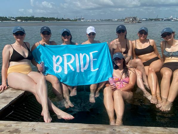 Bachelorette party at a coastal marina: eight women in swimsuits and matching caps sit on a wooden dock with their feet in the water, holding a bright turquoise banner reading BRIDE under a sunny, partly cloudy sky with boats in the background.
