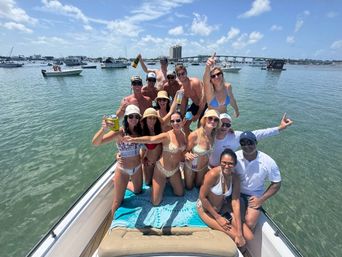 Boat party with a smiling group in swimsuits on the bow of a boat in a sunny coastal bay, holding drinks with anchored boats and a bridge visible on the horizon