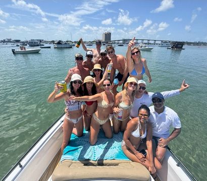 Boat party with a smiling group in swimsuits on the bow of a boat in a sunny coastal bay, holding drinks with anchored boats and a bridge visible on the horizon