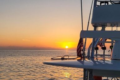 Golden-orange ocean sunset viewed from a boat deck, with an American flag and fishing rod holders silhouetted against calm water and sky.