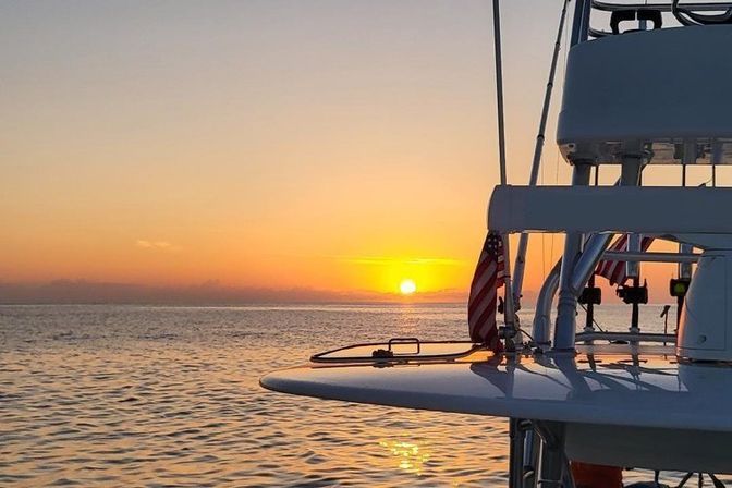 Golden-orange ocean sunset viewed from a boat deck, with an American flag and fishing rod holders silhouetted against calm water and sky.