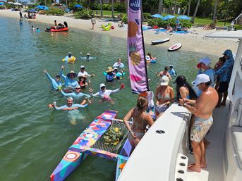 Sunlit boat-and-beach party with swimmers waving around a colorful floral float topped with pineapples, paddleboards and umbrellas along a sandy tropical shore.