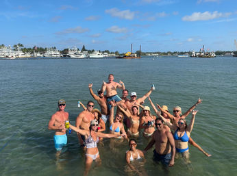 About 15 people in swimsuits cheering with drinks in shallow coastal water by a yacht-filled marina under a sunny blue sky