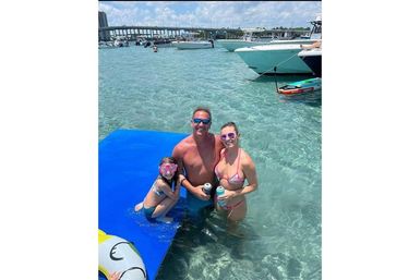 Family of three on a blue floating mat at a coastal sandbar in clear turquoise shallow water, surrounded by anchored boats and a bridge; adults holding canned drinks and child wearing pink snorkel mask.