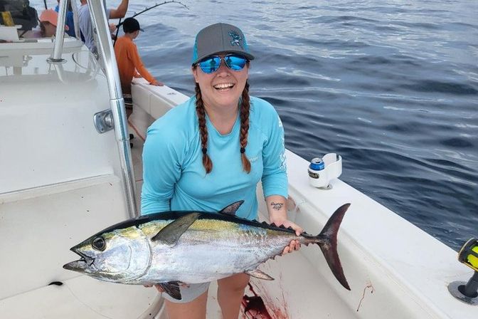 Smiling angler in blue long-sleeve shirt and cap holding a large yellowfin tuna on a fishing boat deck with the open ocean in the background — offshore deep-sea fishing catch.