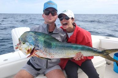 Two excited anglers on a white boat proudly holding a large iridescent mahi‑mahi (dolphinfish) after a deep‑sea fishing catch, open ocean and cloudy sky in the background.