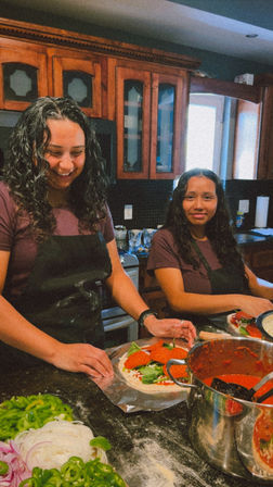 Two people in a home kitchen making homemade pepperoni pizzas on a flour-dusted counter, with a pot of tomato sauce and bowls of sliced peppers and onions nearby.