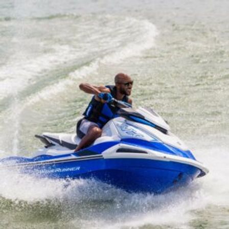Person on a blue-and-white jet ski slicing through choppy lake water, wearing a life jacket and sunglasses as spray and wake trail behind—energetic summer watersports shot.