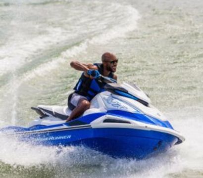 Person on a blue-and-white jet ski slicing through choppy lake water, wearing a life jacket and sunglasses as spray and wake trail behind—energetic summer watersports shot.