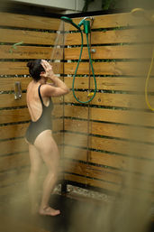 Back view of a person in a black one‑piece rinsing hair under a green outdoor showerhead mounted on a wooden slat privacy fence, barefoot on a dark shower mat.