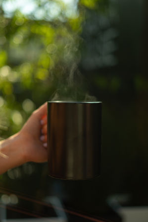 Steaming black mug held by a hand, warm vapor curling upward against a sunlit green bokeh background.