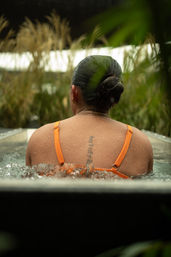 Rear view of a person in an orange swimsuit sitting in an outdoor hot tub, water bubbling around their shoulders, droplets on skin, hair in a neat bun and a vertical script tattoo along the upper spine, framed by tall grasses and greenery.