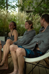 Three friends wrapped in gray towels relaxing and chatting on white chairs at an outdoor poolside deck with tall ornamental grasses in the background.