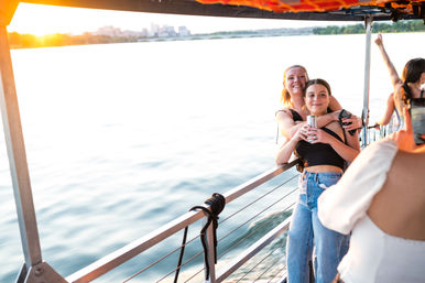 Two friends hugging and posing with a canned drink on the railing of a sunset boat cruise, calm water below and a distant city skyline on the horizon.