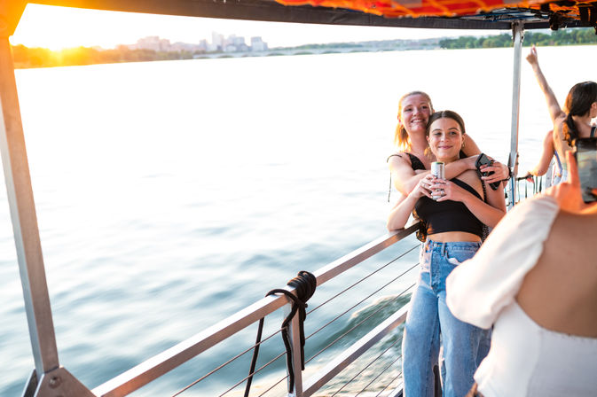 Two friends hugging and posing with a canned drink on the railing of a sunset boat cruise, calm water below and a distant city skyline on the horizon.