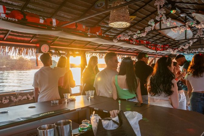 Tiki‑roofed sunset boat party on the water, casual crowd gathered at a deck bar with drinks as warm golden light reflects across the water.