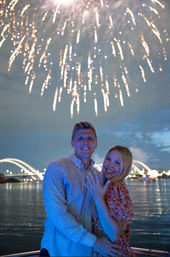 Smiling couple on a nighttime riverside boat, woman showing her engagement ring as golden fireworks burst over illuminated arched bridges and calm water.