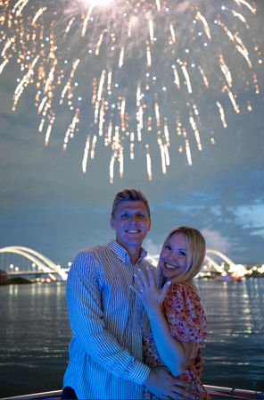 Smiling couple on a nighttime riverside boat, woman showing her engagement ring as golden fireworks burst over illuminated arched bridges and calm water.