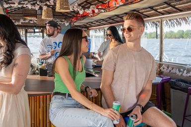 Friends chatting at a tiki-style boat bar on a sunny river cruise, woman in a green tank top and man in sunglasses sipping canned drinks