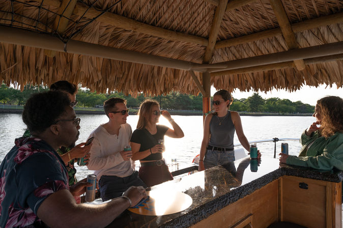 Group of friends socializing under a thatched tiki-style bar on a boat at sunset, holding canned drinks with a tree-lined lake shore in the background